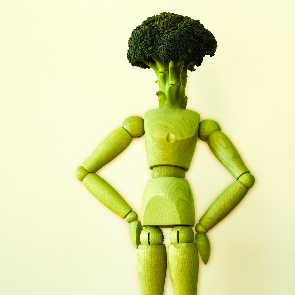 Wooden manikin standing in front of a yellow background. The manikin is green and its head is replaced by a broccoli.