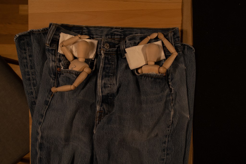 Pair of manikins, seemingly sleeping, tucked each in the pockets of a pair of jeans, using folded tissues as pillows.