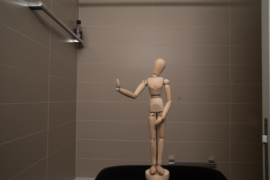 Wooden manikin looking puzzled, standing in front of bathroom tiles.