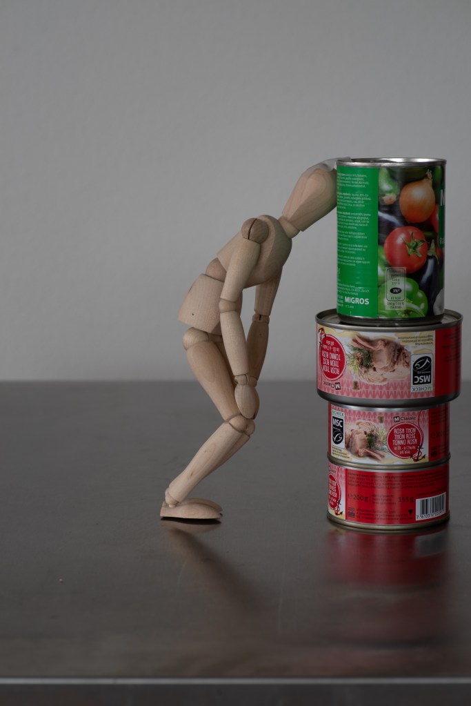 Wooden manikin slouching in front of a white wall, holding thanks to a pile of tin cans supporting its head.