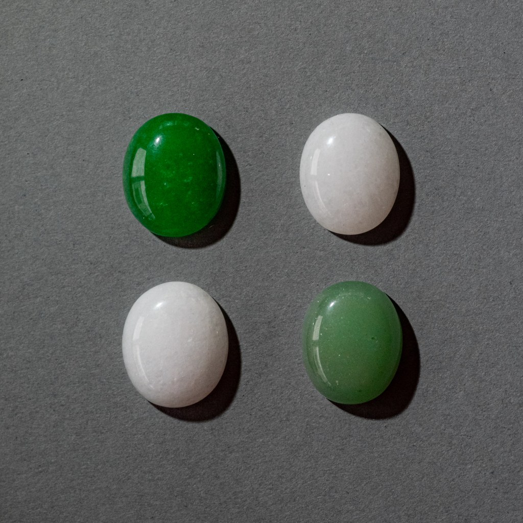 Four small oval gemstones, aligned on the four corners of a square, on a grey cardboard background. The upper right and lower left stones are white, the two others are dark green and light green.