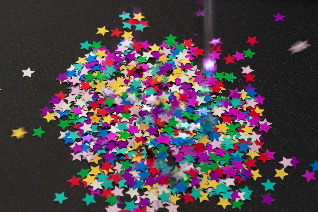 A bunch of colorful star confetti gathered on a black surface; some more confetti are falling.