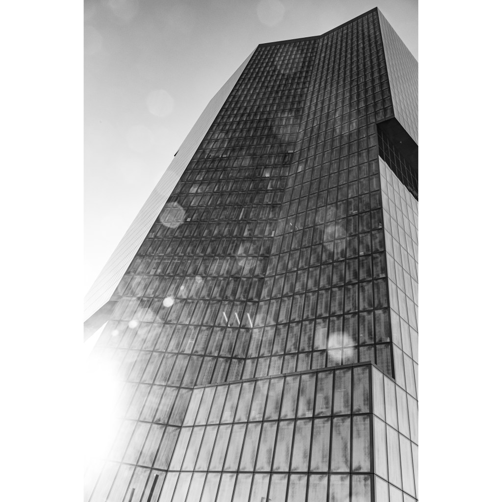 Black and white photography of the Prime Tower in Zürich. The Sun visible on the lower left side of the picture distributes a large amount of bubble-like lens flares all around the image.