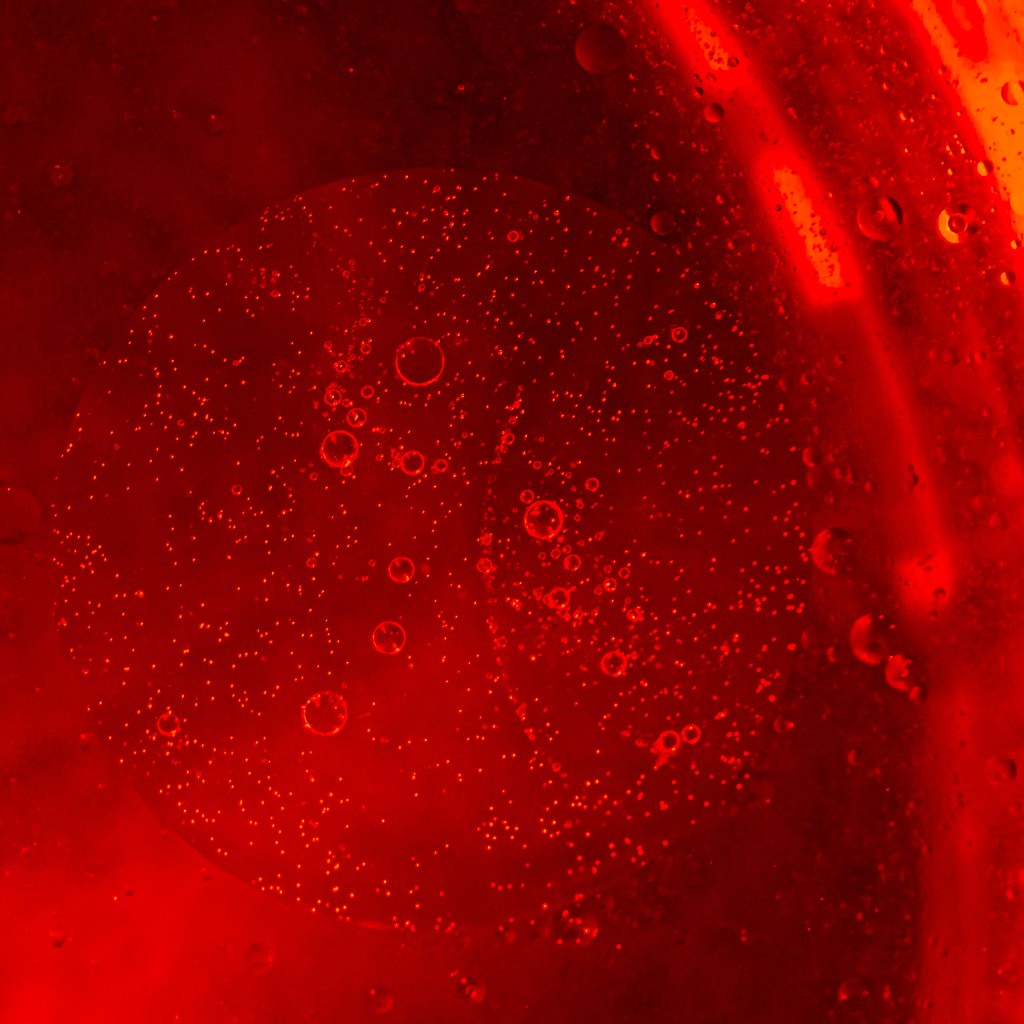 Abstract photography in red tones. There's a larger circle that takes almost the whole square picture; that circle contains numerous small bubbles. Outside of the circle, there's a larger circular arc on the right, also scattered with bubbles, that fades to orange/yellow tones.