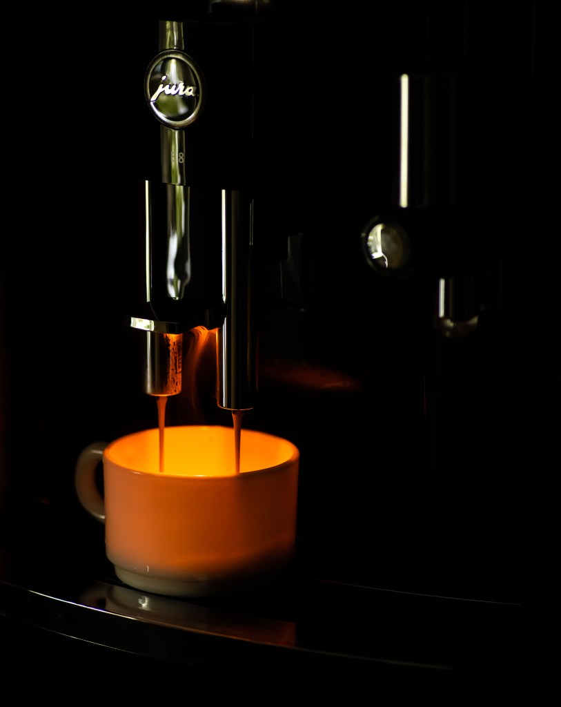 A low-key picture of an espresso machine making an espresso. The white cup in the bottom is lit in orange; a small trail of vapor is visible on top.