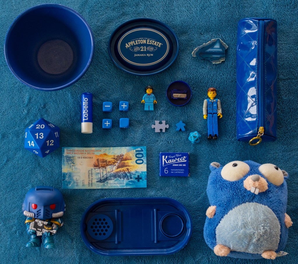 A bunch of blue stuff on a blue blanket. From left to right and top to bottom: a bowl, a rum metallic cover "Appleton Estate Jamaica Rum", a piece of agate, a pencil case, a d20 showing number 13, a Labello stick, four Fate dice showing a minus, two plusses and a blank, a LEGO woman conductor minifig, a pencil sharpener, a LEGO Technic figurine, the back of a jigsaw piece, a Meeple, a Star Trek D20 showing a Star Trek logo, a 100CHF bill, a box of Kaweco blue ink, a Space Marine Funko Pop toy, a plastic box lid, and a stupid looking gopher Go mascot plush.