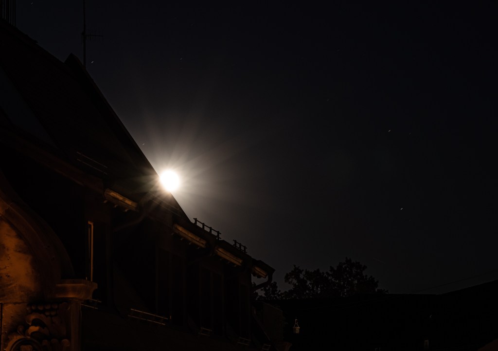 Overexposed moon appearing behind a building. The moon seems to throw straight rays in all directions.