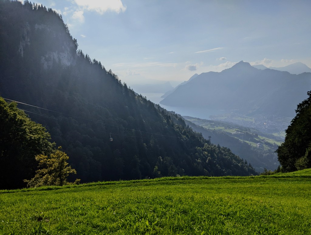 Landscape picture. The foreground is green grass; then there's a mountain with a small cable car and sun rays filtering through trees; the background shows the side of a lake ans more mountains in the distance. 