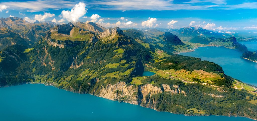 View over blue/turquoise lakes and green lush mountains with a few clouds and a very blue sky.