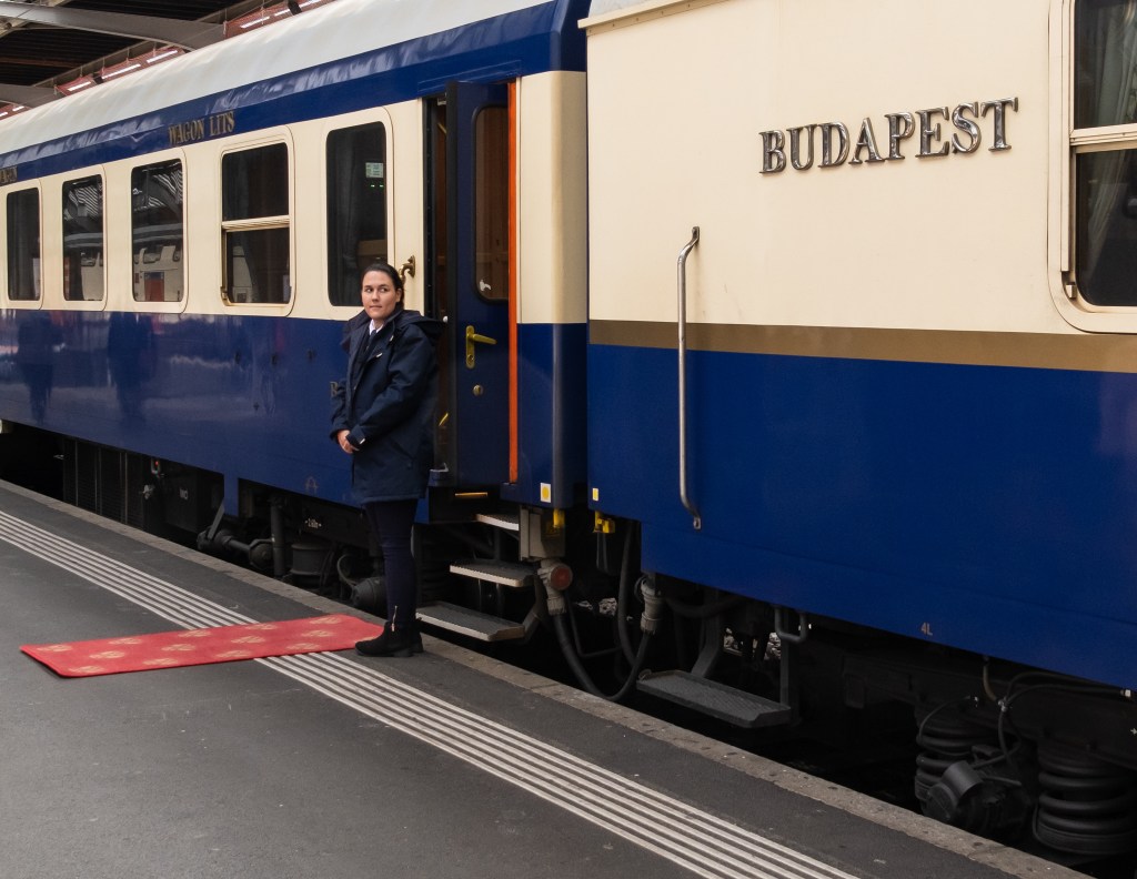 A train attendant standing in front of the open door of an older train. The train beige, blue and golden; the letters BUDAPEST are visible on its side.