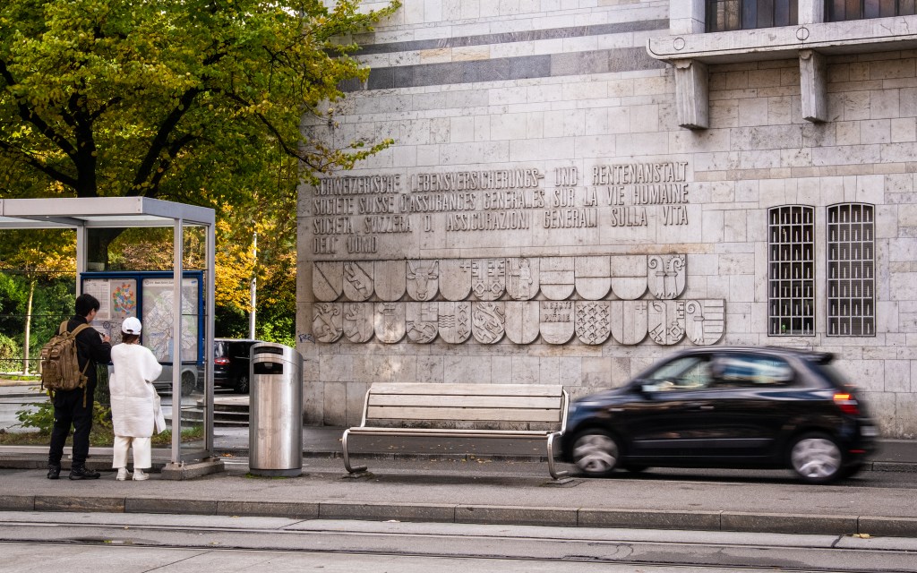 Facade of SwissLife, showing the letters "Swiss society of general human life insurance" in German, French and Italian, with all Swiss cantons flags below, carved in stone. In front of the wall, a small black car is passing quickly, and two people are looking at tram times at the tram stop. A large green and yellow tree is visible in the background of the image.
