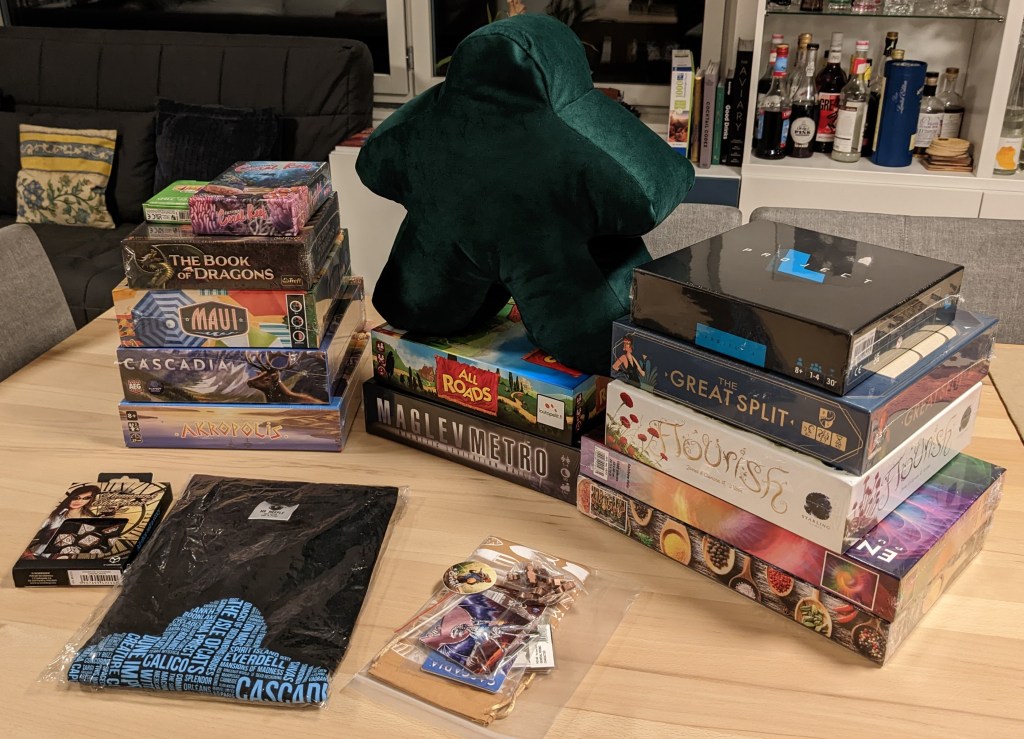 Loot from Essen SPIEL - multiple game boxes (Coral Reef, The Book of Dragons, Maui, Cascadia, Akropolis,All Roads, Maglev Metro, Project L, The Great Split, Flourish), a couple of jigsaw puzzles, a tshirt and a dark green meeple cushion.