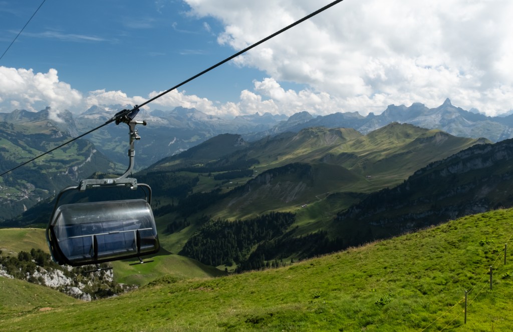 A chairlift cabin next to a mountain landscape. The sky is blue with some clouds, the mountains are visible at some distance.