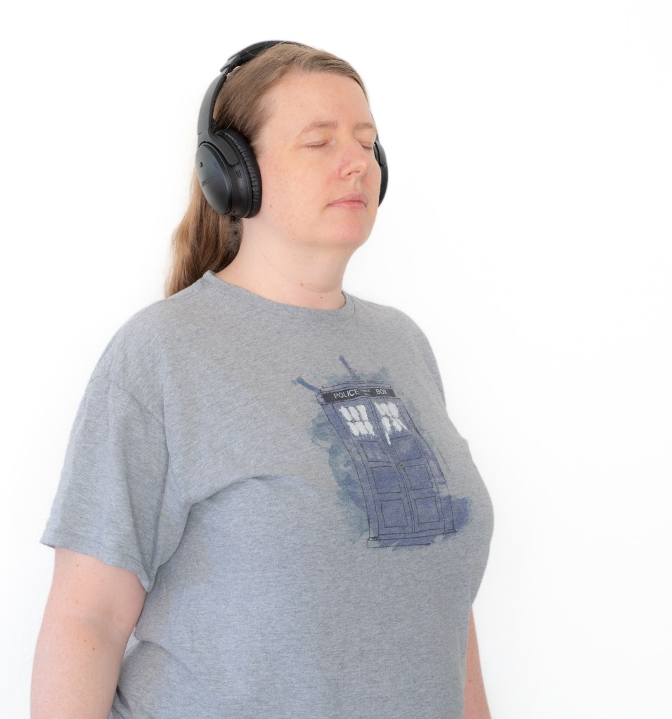 A woman wearing a large headset and a Doctor Who blue TARDIS t-shirt, looking calm.