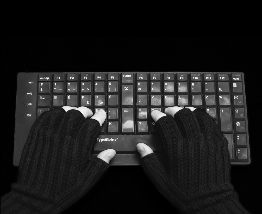 Black and white picture of hands in fingerless gloves over a Typematrix BÉPO keyboard, on a black background. The keyboard shows some wear and tear.