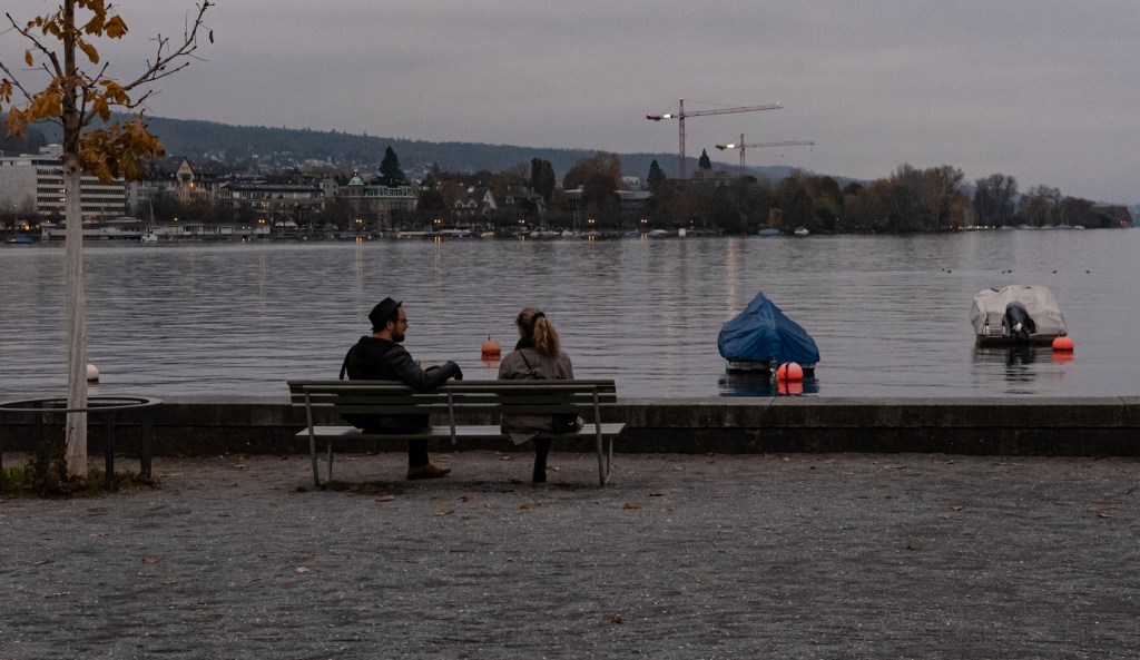 A man wearing a hat and a woman sitting on a bench facing the lake. Two covered boats with red buoys are visible on the lake; the city and some trees are visible in the background of the lake. Two cranes surplomb the city.