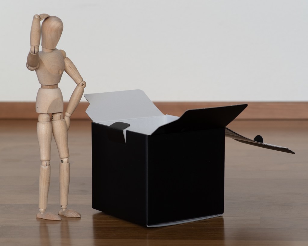 A wooden manikin, looking perplexed, in front of an open black box.