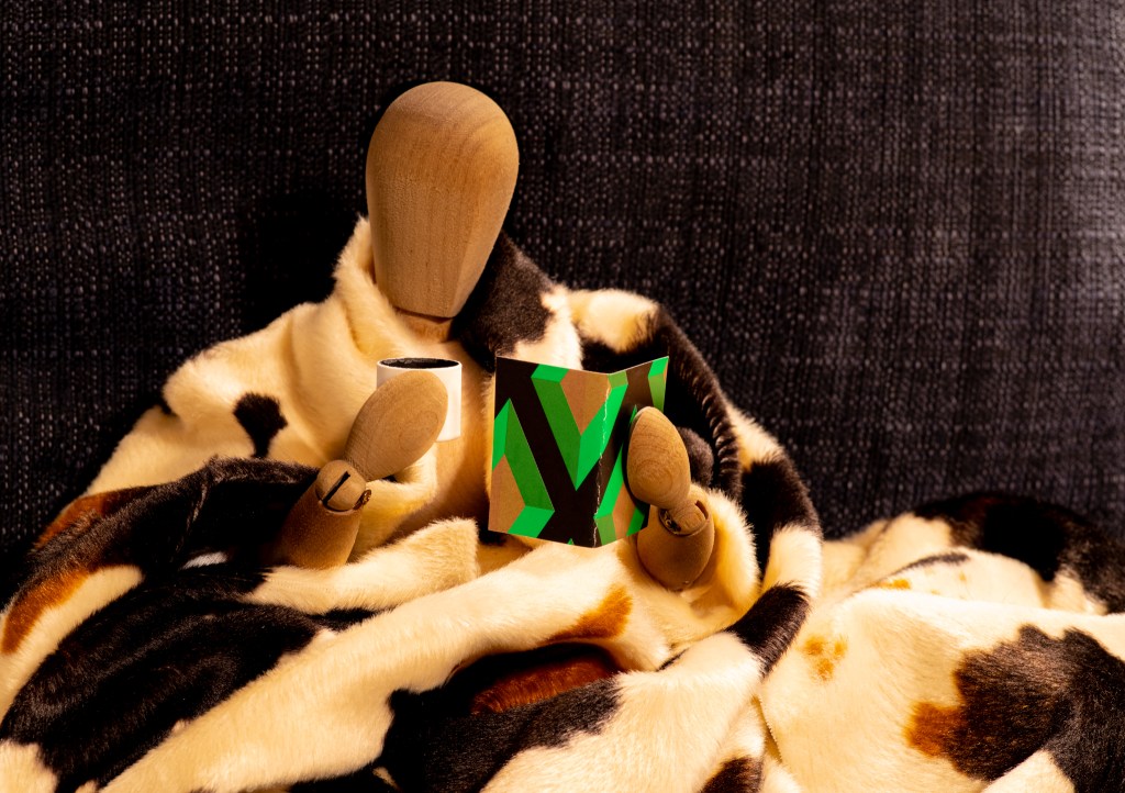 A wooden manikin looking cozy in a cow-spots-patterned blanket, holding a mug in one hand and a green book in the other.