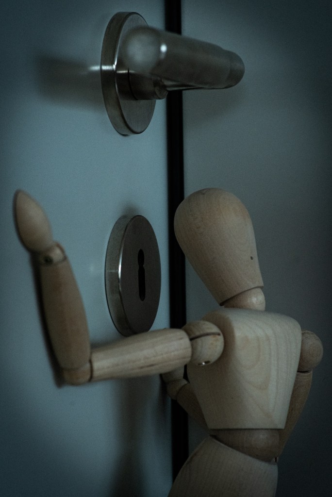 A wooden manikin seemingly spying at something through a keyhole, processed with vignetting, grain and blue tones.