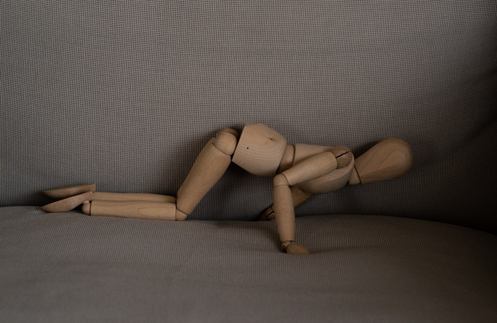 A wooden manikin on all fours, with its arm stuck between the cushions of a sofa, probably looking for its keys.