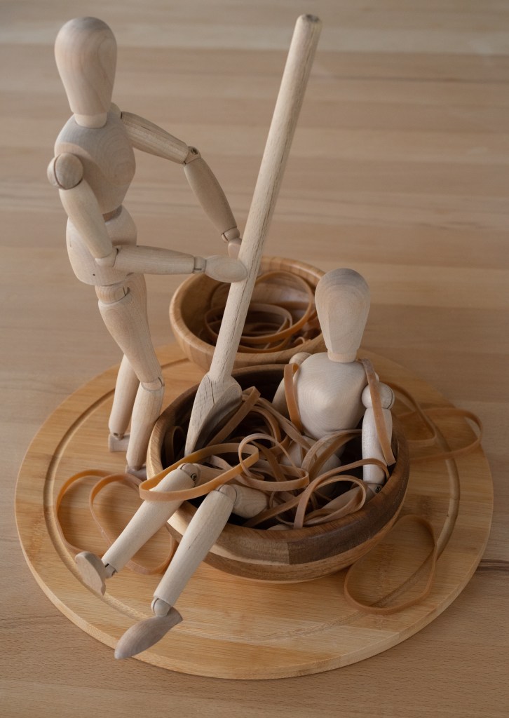 Two wooden manikins. One is sitting in a wooden bowl filled with elastic bands; the other is stirring the bowl with a wooden spoon. 