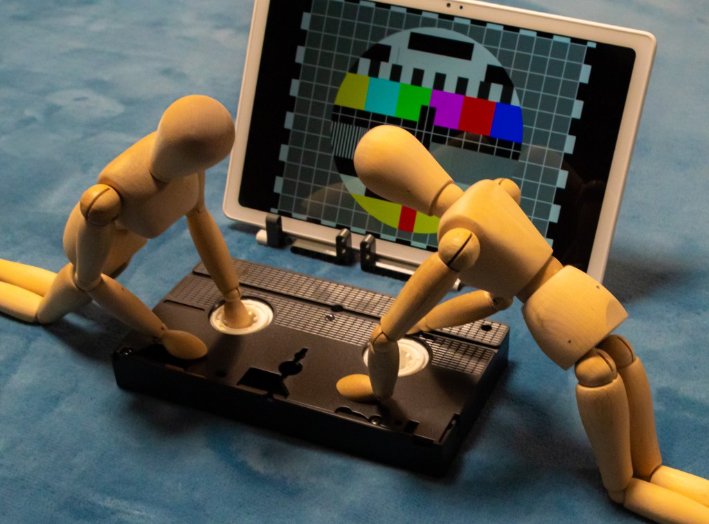 Two wooden manikins on all fours, busy rewinding a VHS tape in front of a TV showing an "end of programmes" test card.