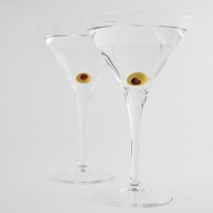 Two martini glasses with a clear liquid and an olive in each, over a seamless grey/white background
