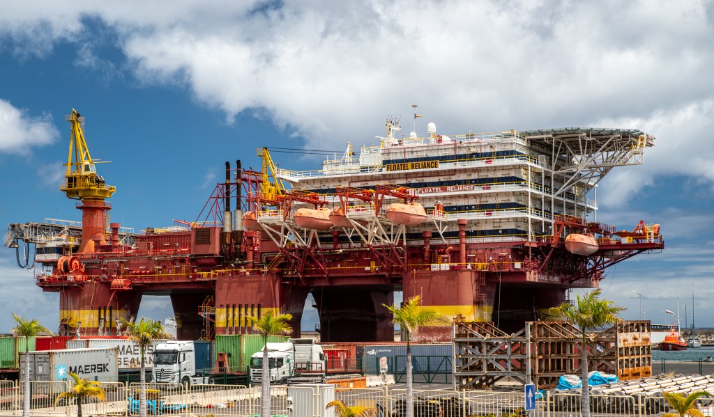 A large accomodation barge, presenting as a small building on top of a large platform on top of six large feet. The platform and the feet are red, offering a stark contrast with the blue sky.