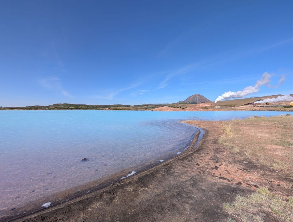 The shore of a very blue lake with, in the background, a triangular mountain and some buildings emitting a thick white smoke