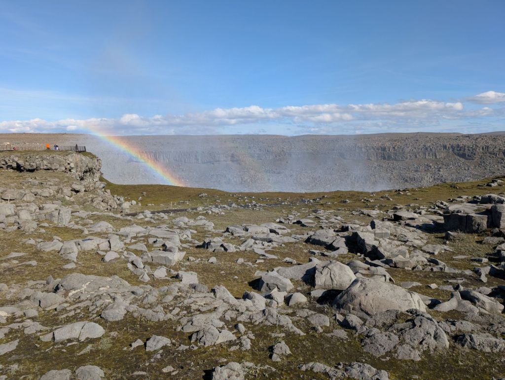 A field of rocks ending on a hole, with another rocky wall visible in the background. A rainbow is jumping from the hole, along with a visible mist. No water is visible apart from the mist.