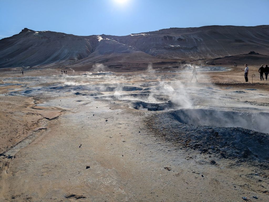 Multiple small grey smoking craters on a flat grey and yellow ground, with mountains in the background