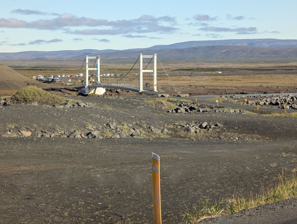 A white suspension bridge over a river, with a line of cars waiting behind it. The bridge is closed for works.