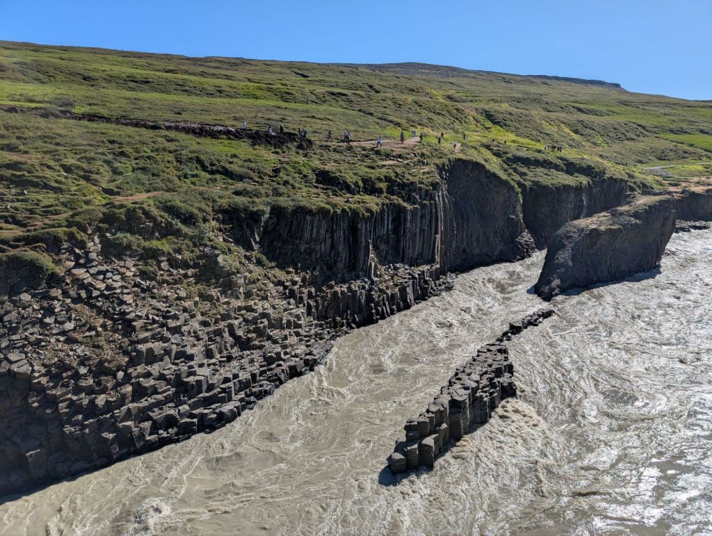 A powerful grey river next to a basalt columns wall; there’s two additional basalt islands in the middle of the river, including one that looks like a gigantic stone alligator