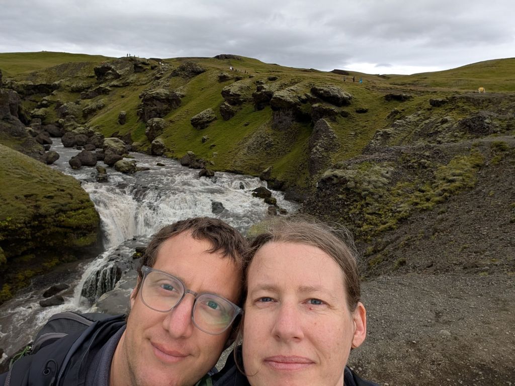 A selfie of a man and a woman smiling awkwardly in front of a waterfall