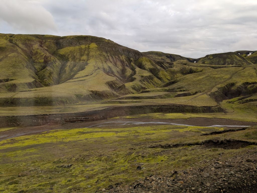 Some green and black mountains with pleasing curvy patterns, and a small stream of water in the foreground