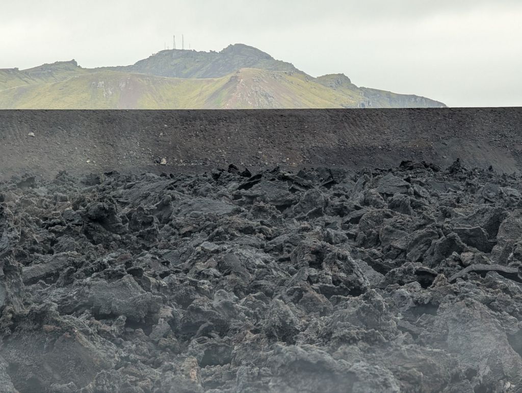 A black lava field behind a dark gravel barrier, with a mountain in the background