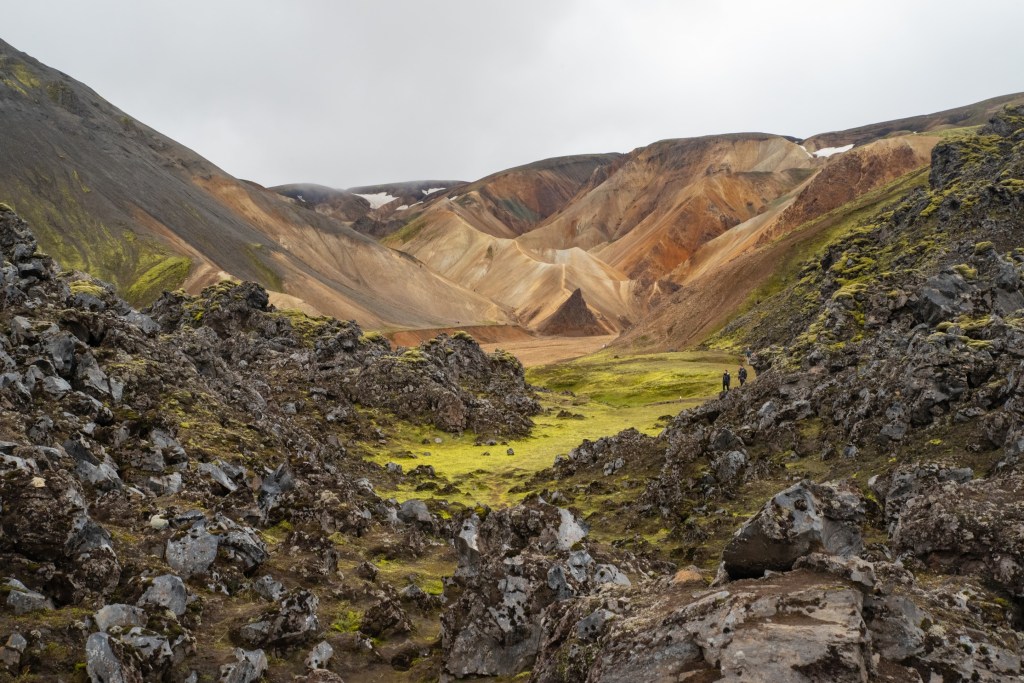 Brown and red mountains, with a clearly visible ridge/path in the middle; in the foreground, dark lava stones and some moss.