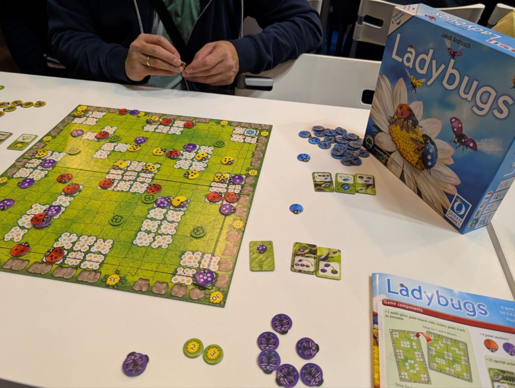 A green board with squares, and fields of daisies taking over some of the squares. There's a bunch of ladybugs tokens with varying numbers of dots on their back in various places on the board.
