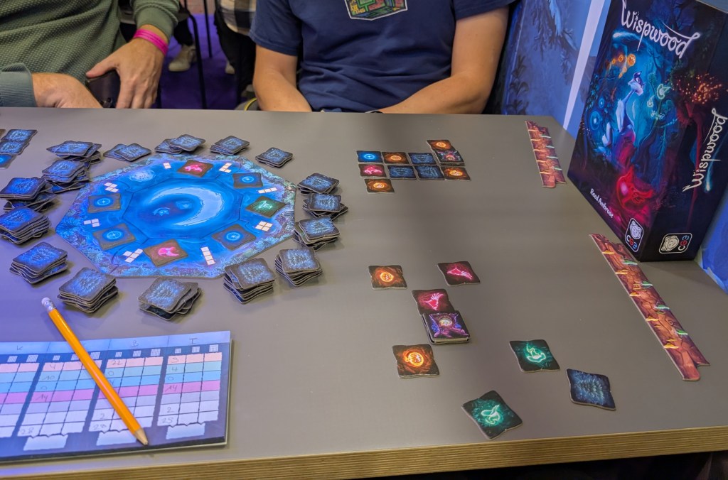 A wheel with square tiles representing firs on the back and various colorful wisps/spirits on the front side, with polyominos on the wheel between each pair of tiles. Players assemble these tiles in front of them in a grid.