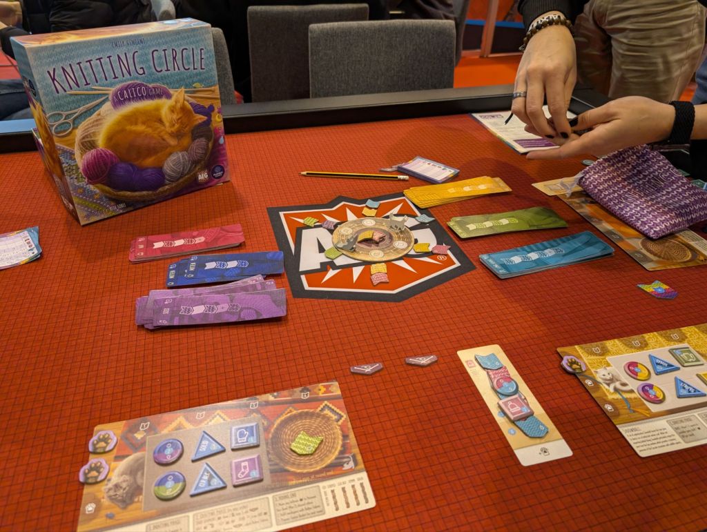 Boards with buttons and yarn, a wheel with two cat tokens and yarn samples around them, and knitting pattern-describing cards.
