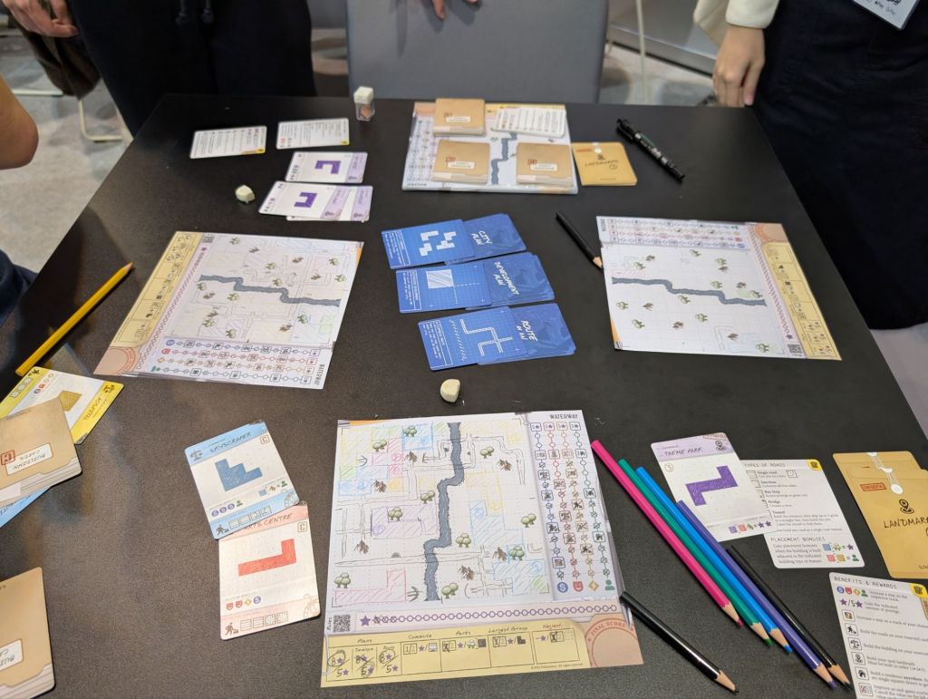 Cards with polyominos and sheets of paper with a lot of roads and polyominos drawn on them. There's a set of colored pencils next to the sheet of paper.