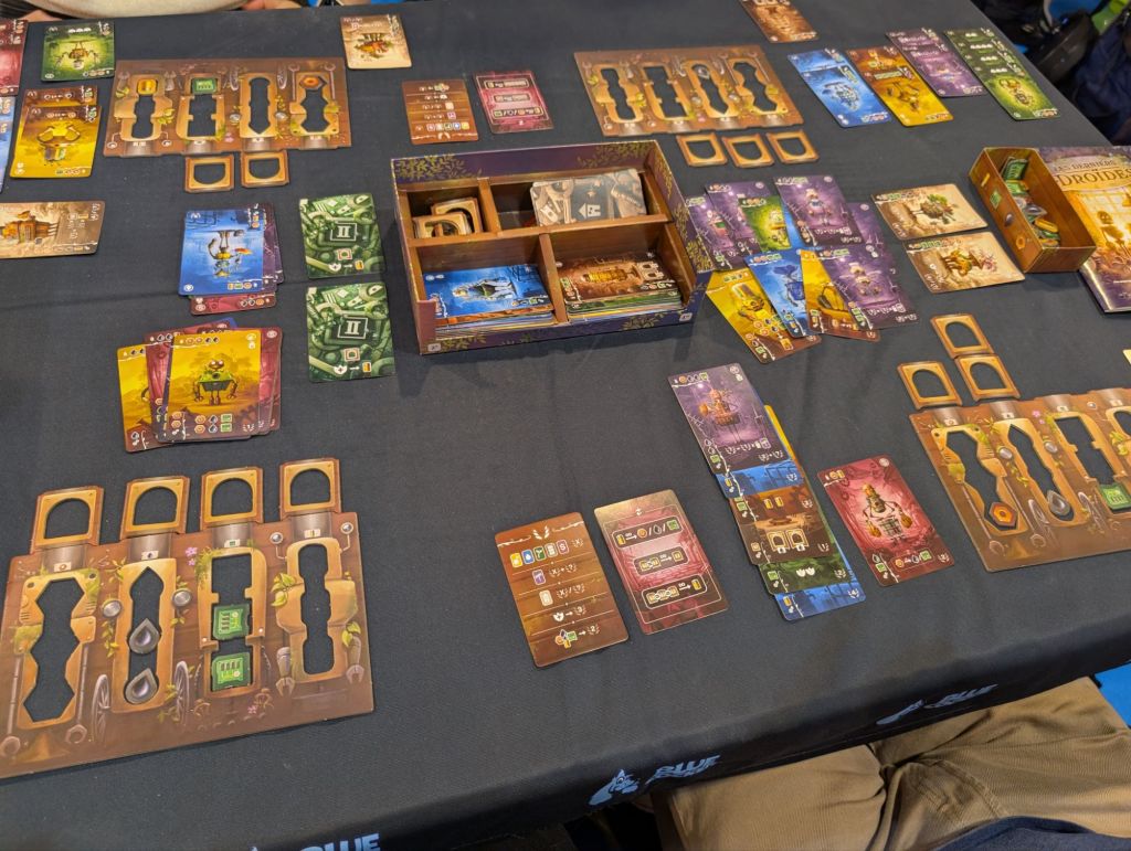 A central tray containing two columns of cards, and player boards with resources (oil, cogs, circuits, batteries). There are a lot of colorful cards with robots on the table.