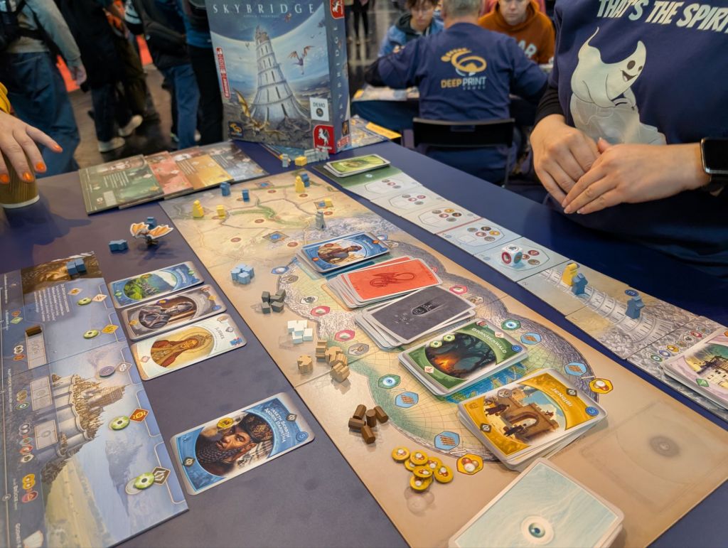 A central board with cards of different colors, a player board with resources and cards on top; a board representing a large bridge/tower being built.